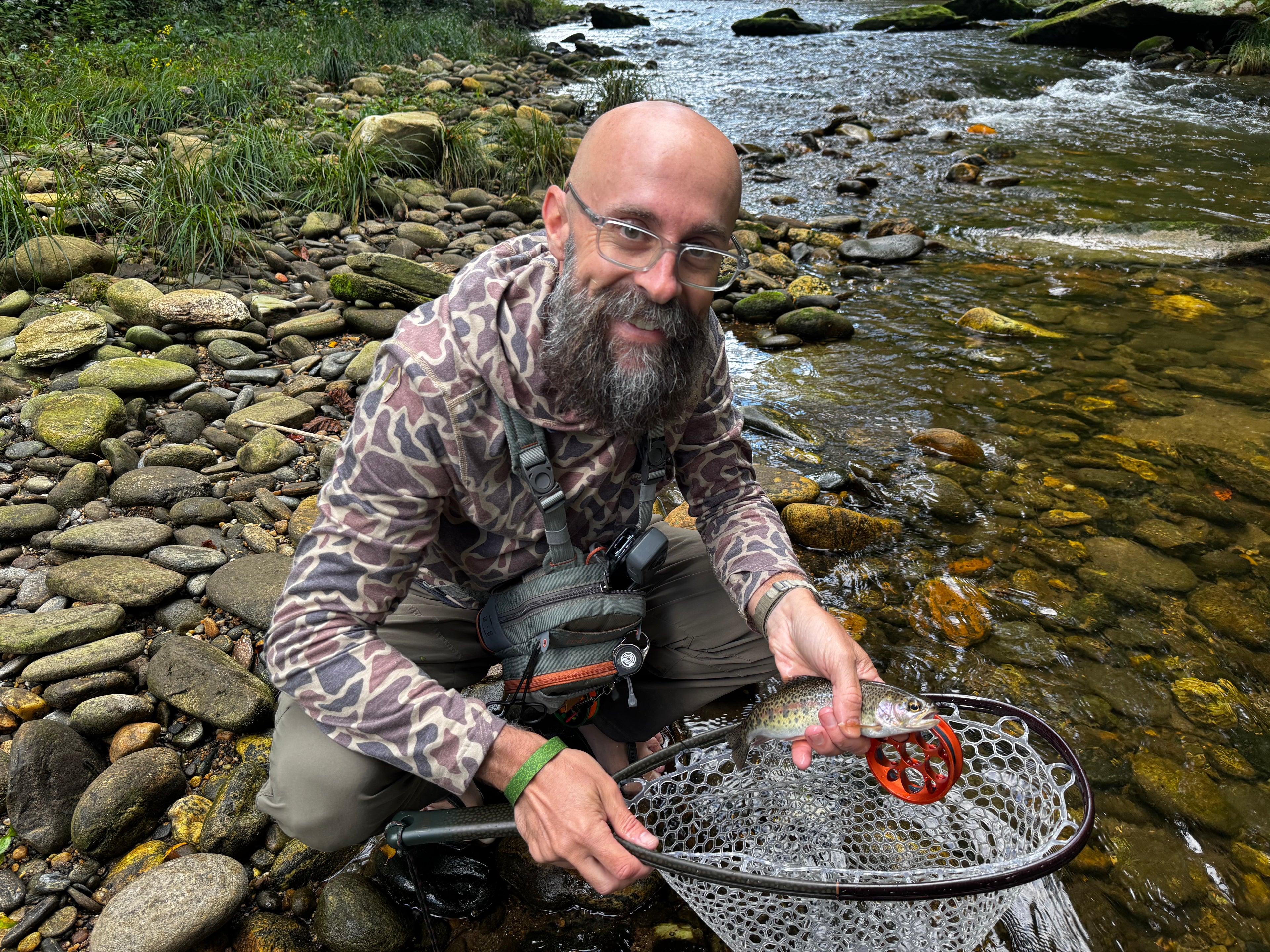 Man fishing by a stream, holding a net with a fish inside.